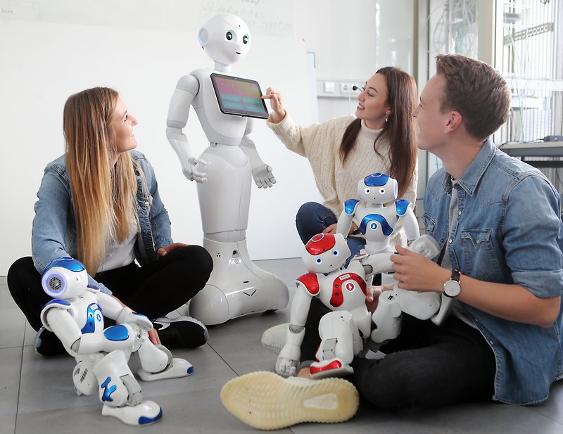 Drei Personen sitzen vor einem großen Roboter, ein weiterer kleiner Roboter sitzt am Boden und zwei weitere kleine Roboter sitzen auf einer der Personen.