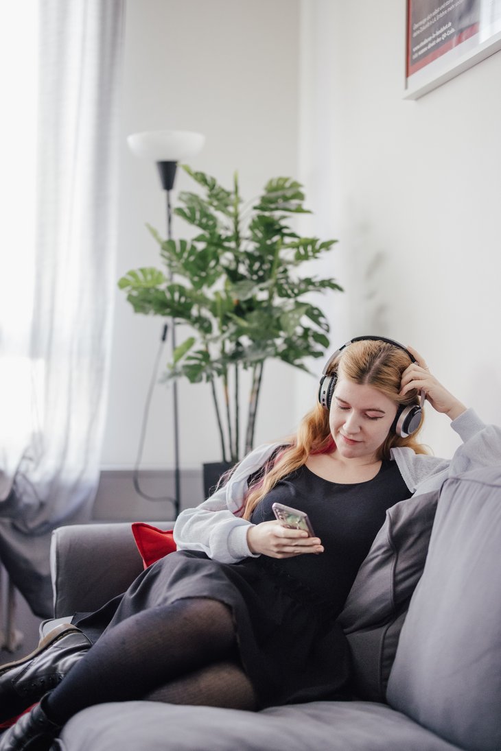 Eine Person sitzt entspannt auf einem grauen Sofa, trägt Kopfhörer und schaut auf ihr Smartphone. Im Hintergrund steht eine Pflanze und ein Stehlampe sorgt für sanftes Licht.