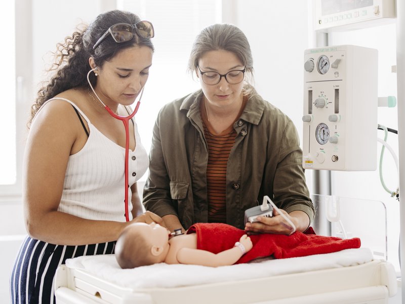Zwei Frauen stehen an einer Neugeborenen-Reanimationseinheit in welchem eine Babypuppe liegt.