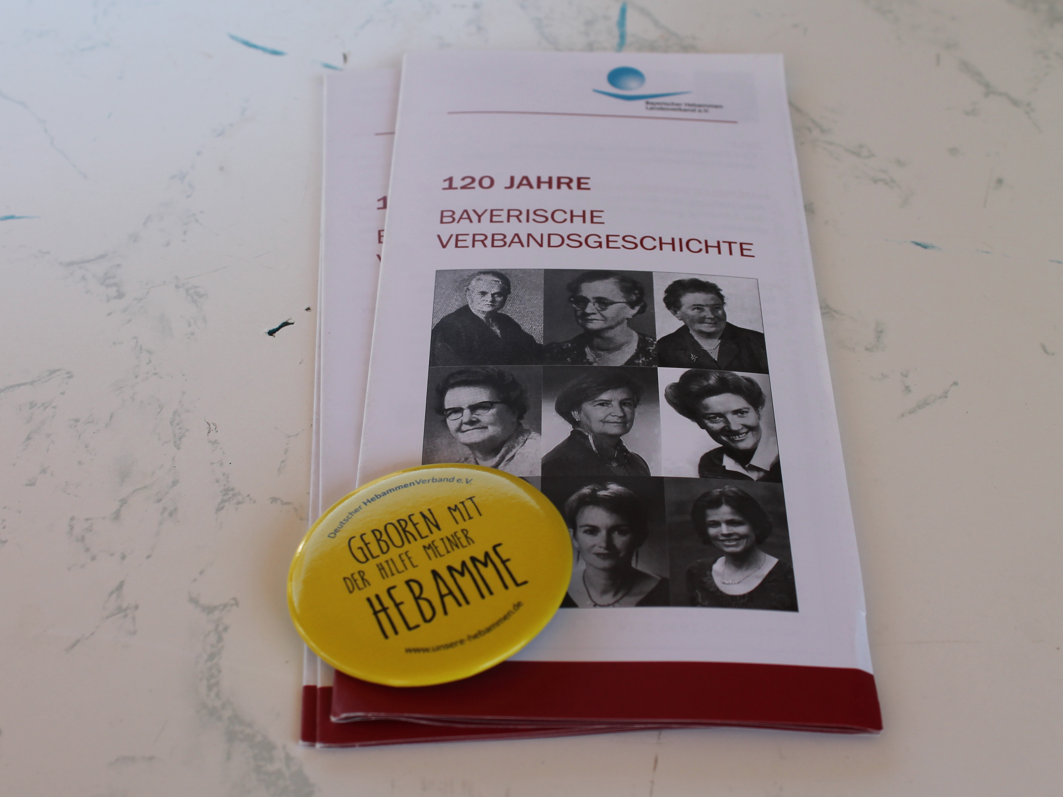 Man sieht einen Flyer und einen Button zur Bayerischen Verbandsgeschichte der Hebammen.
