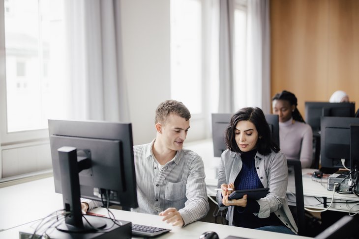 Zwei Personen arbeiten an einem Computer in einem modernen Büro. Eine Person zeigt etwas auf einem Tablet, während die andere aufmerksam zuschaut. Im Hintergrund sind weitere Personen an ihren Computern zu sehen.