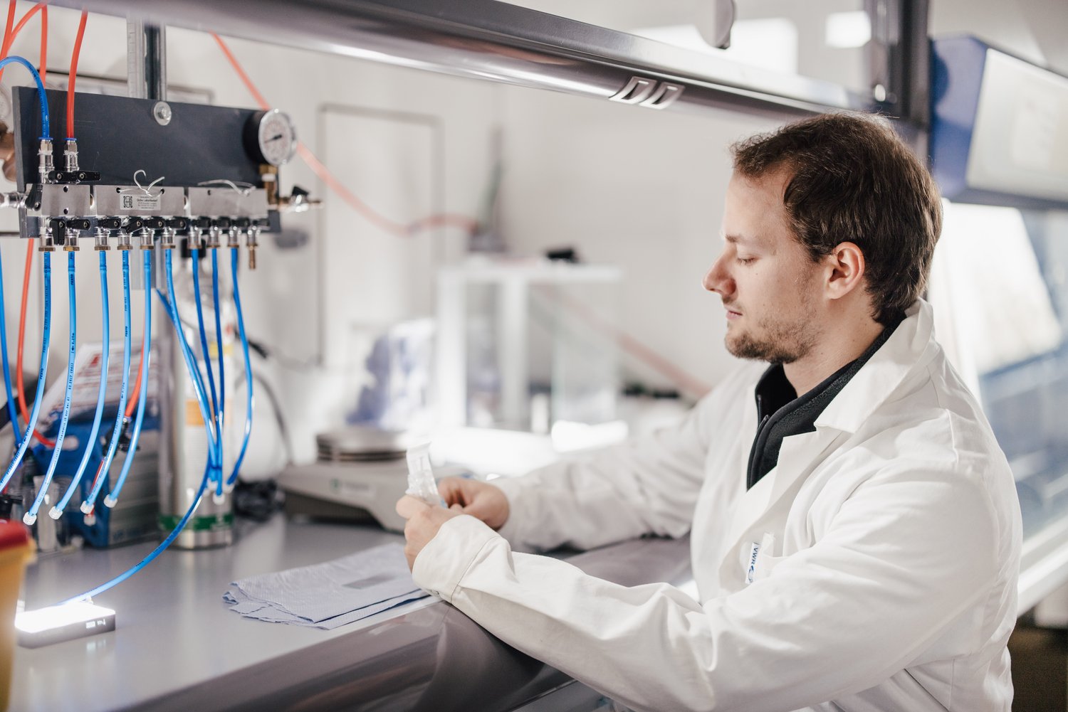 Eine Person in einem weißen Laborkittel sitzt an einem Tisch in einem Labor und betrachtet ein Dokument. Im Hintergrund sind verschiedene Geräte und Schläuche sichtbar, die für wissenschaftliche Arbeiten genutzt werden.