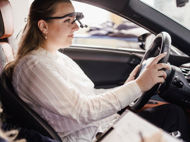 Eine Person sitzt am Steuer eines Fahrzeugs und testet es. Eine weitere Person sitzt daneben und macht Notizen auf einem Clipboard. Beide sind in einem modernen Auto mit einem eleganten Innenraum.