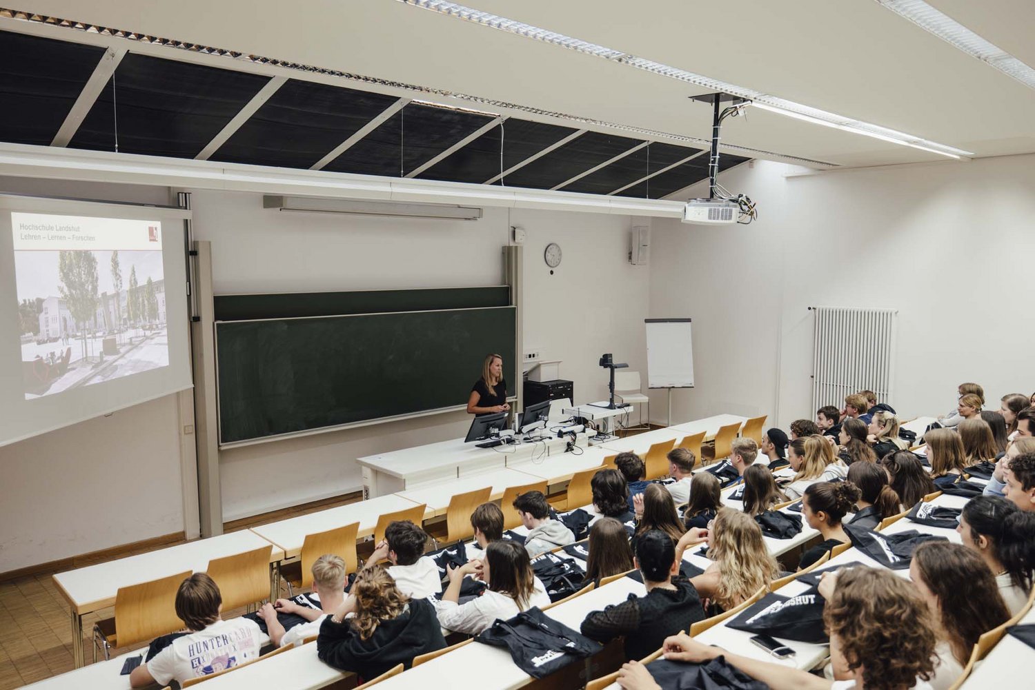 Man sieht mehrere sitzende Personen und eine Person die vorne im Raum an einer Tafel steht.