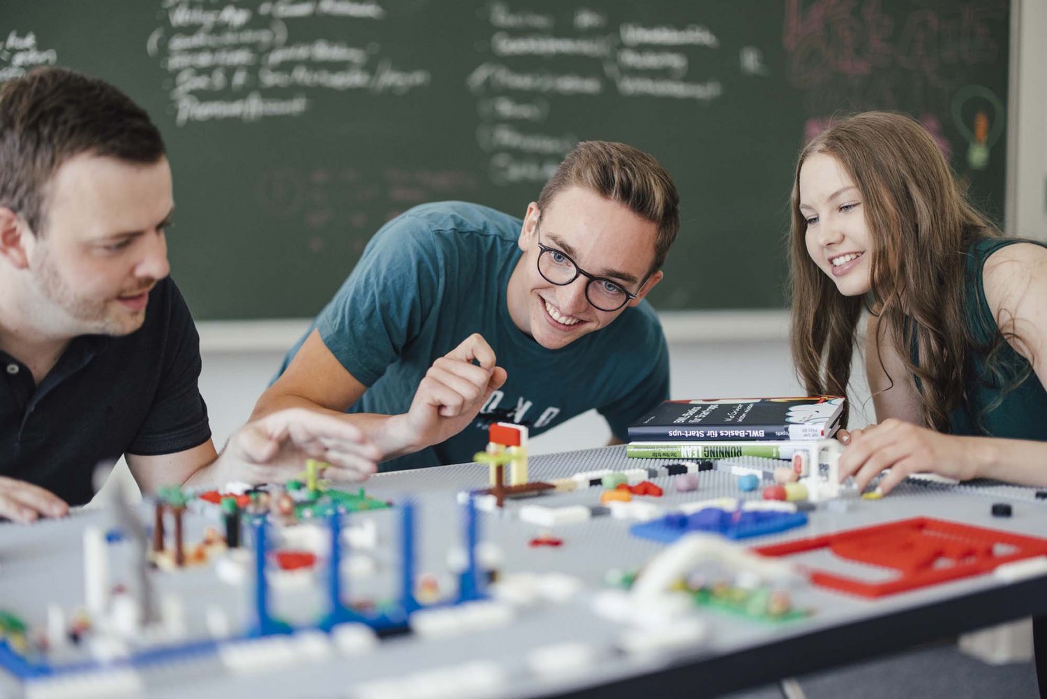 Drei Personen sitzen um einen Tisch herum. Sie lachen. Es sind Bauteile auf dem Tisch zu sehen.