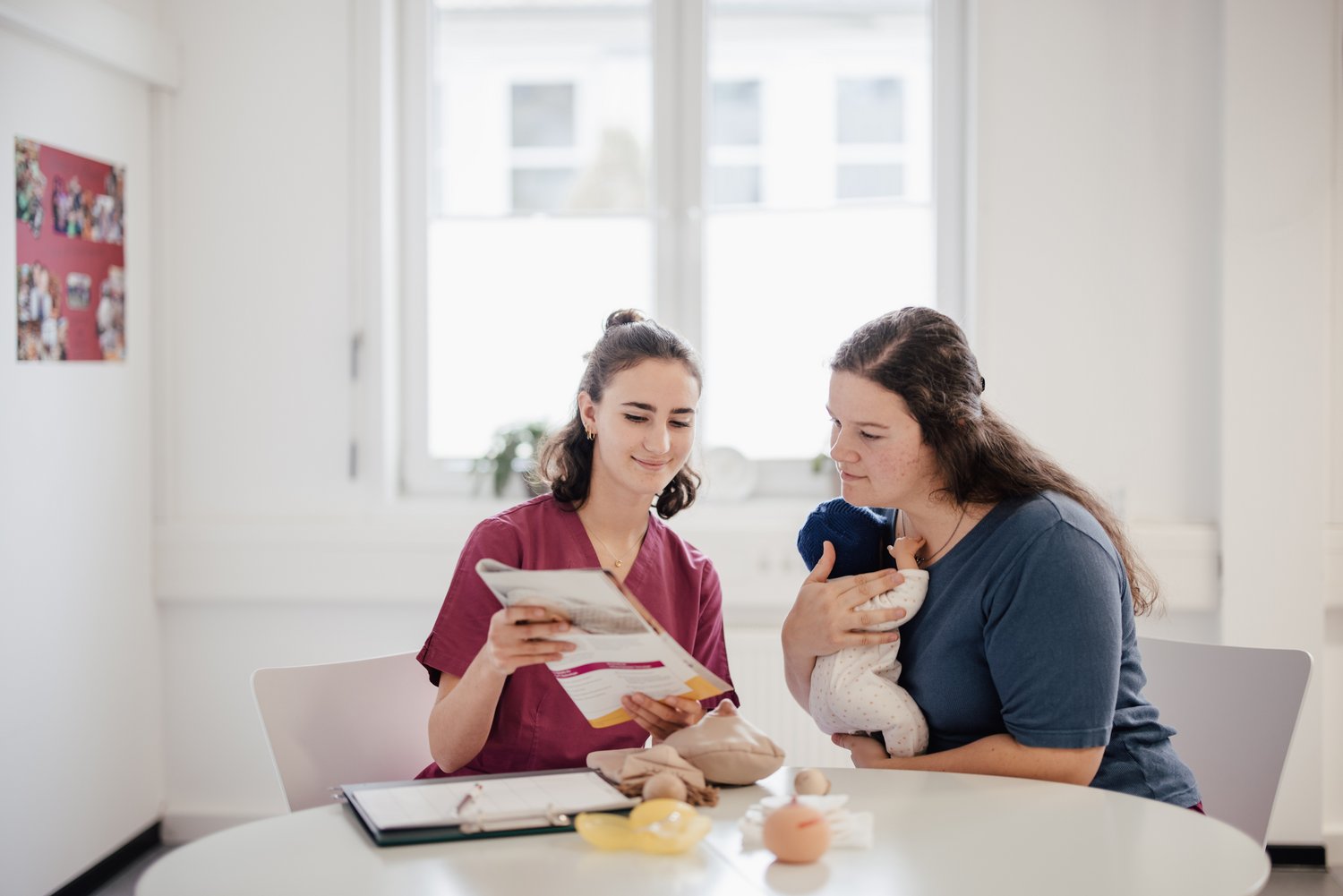 Eine Person in medizinischer Kleidung zeigt einer anderen Person ein Informationsblatt. Letztere hält ein Baby in den Armen. Beide sitzen an einem Tisch in einem hellen Raum mit Fenstern im Hintergrund.