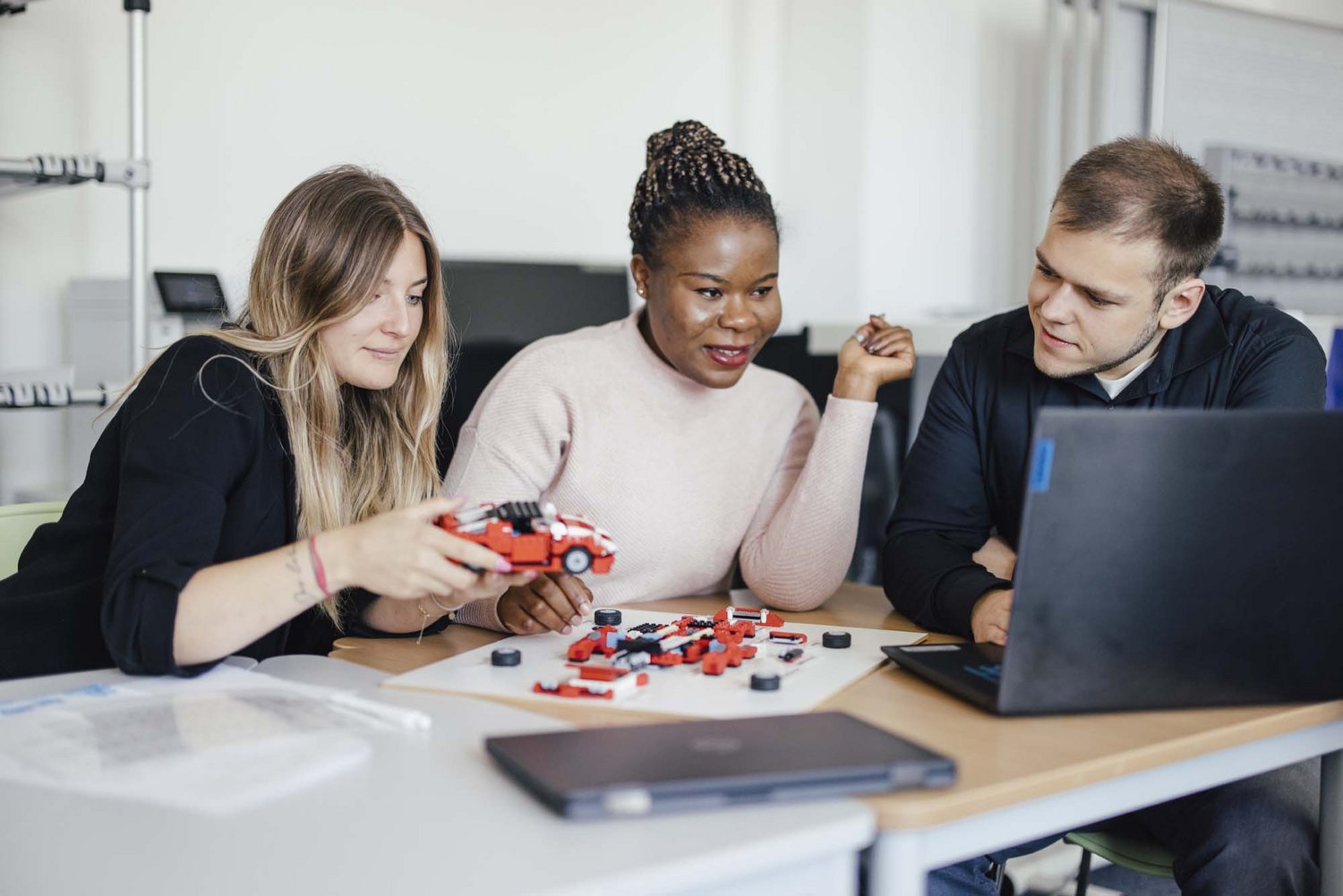 Drei Personen sitzen zusammen an einem Tisch. Die eine Person hält ein selbst zusammengebautes Modellauto in der Hand. Die anderen Personen schauen auf das Auto sowie auf einen Laptop.