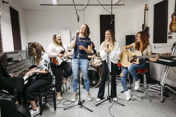 Sechs Personen sind zusehen. Vier davon spielen unterschiedliche Instrument und zwei halten jeweils ein Stehmikrofon in der Hand.