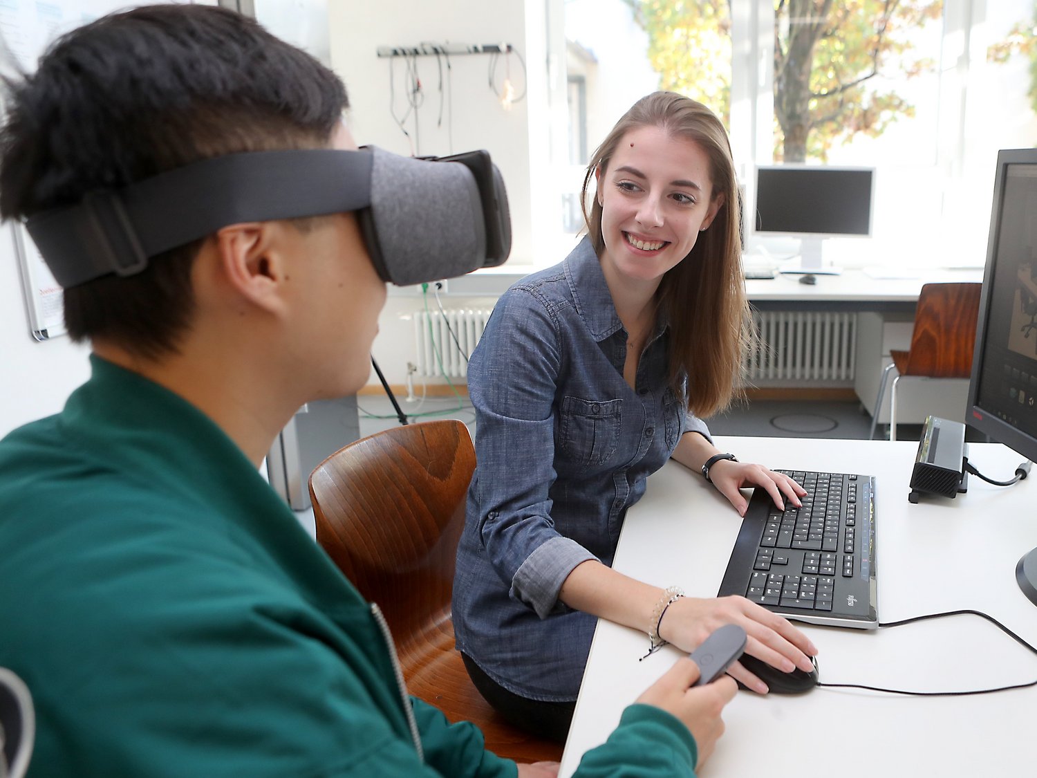 Zwei Personen sitzen vor einem PC. Eine Person trägt eine VR-Brille.