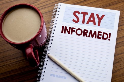 Rote Kaffeetasse auf einem hölzernen Hintergrund, daneben liegt ein weißer Block mit dem Schriftzug "Stay Informed!" in roter Schrift, ein Stift liegt auf dem Block