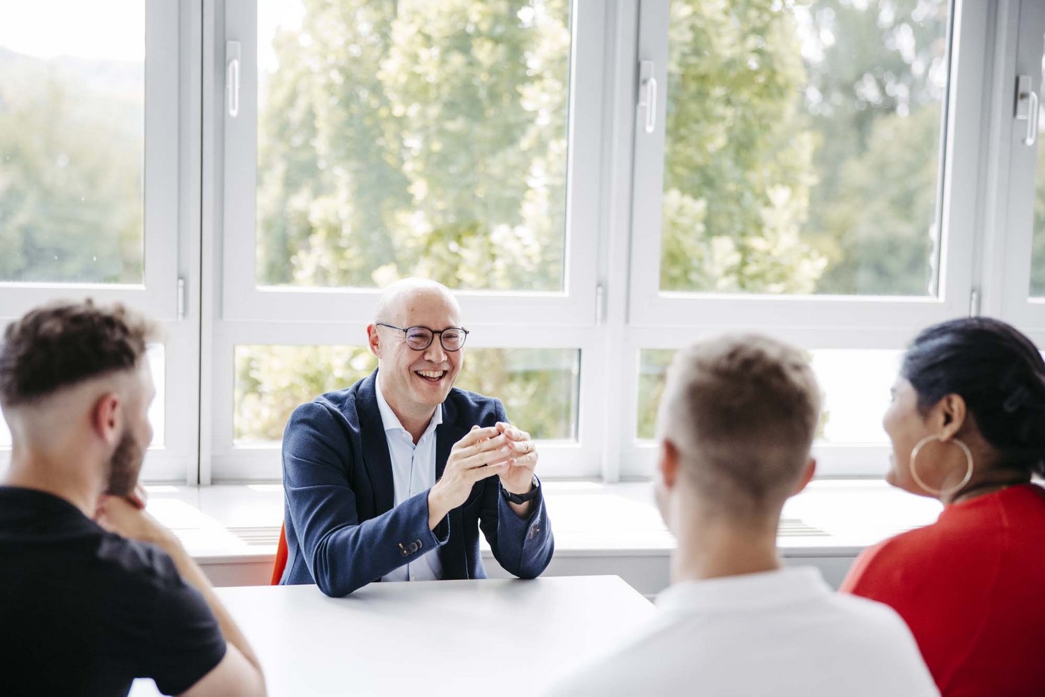 Vier Personen befinden sich einem Raum, drei sitzen nebeneinander, die andere Person sitzt gegenüber. Alle schauen sich gegenseitig an.