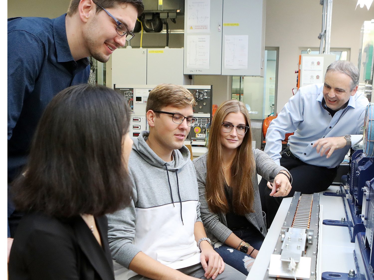 Mehrere Studierende und ein Professor sitzen vor dem Aufbau eines Linearmotors. Sie unterhalten sich.