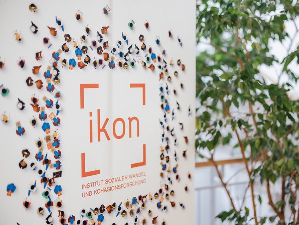 Ein Banner des Instituts für sozialen Wandel und Kohäsionsforschung (ikon) ist umgeben von einer Vielzahl kleiner Figuren, die eine Gemeinschaft symbolisieren. Im Hintergrund ist eine grüne Pflanze sichtbar.