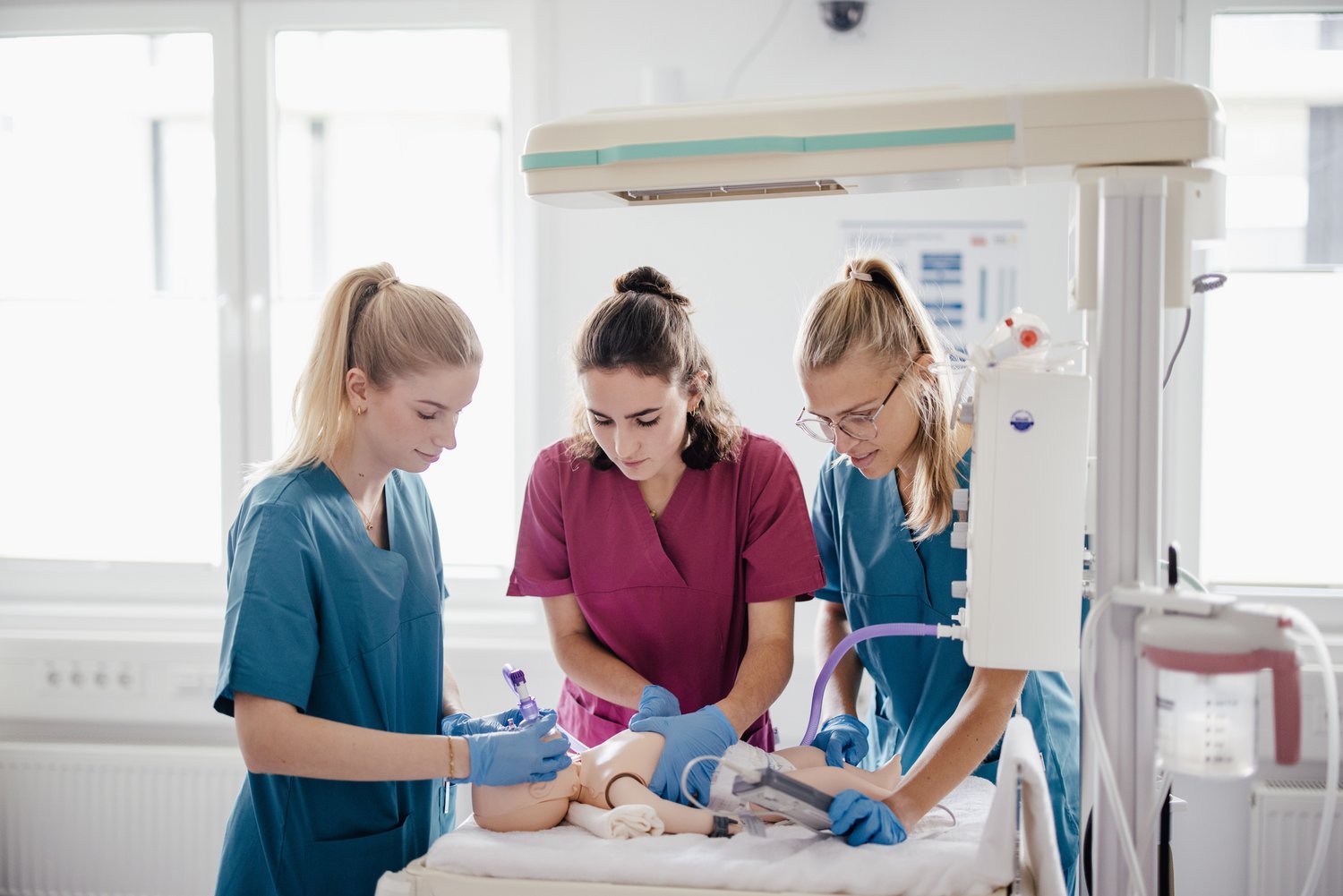 Drei Personen in medizinischer Kleidung arbeiten an einem Simulationsmodell. Eine Person führt eine Injektion durch, während die anderen assistieren. Der Raum ist hell und modern, mit medizinischen Geräten im Hintergrund.