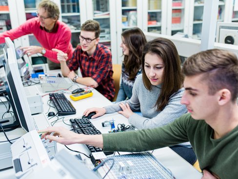 Mehrere Studierende sitzen an Arbeitsplätzen zum Messen und Testen. Ein Dozierender erklärt etwas.