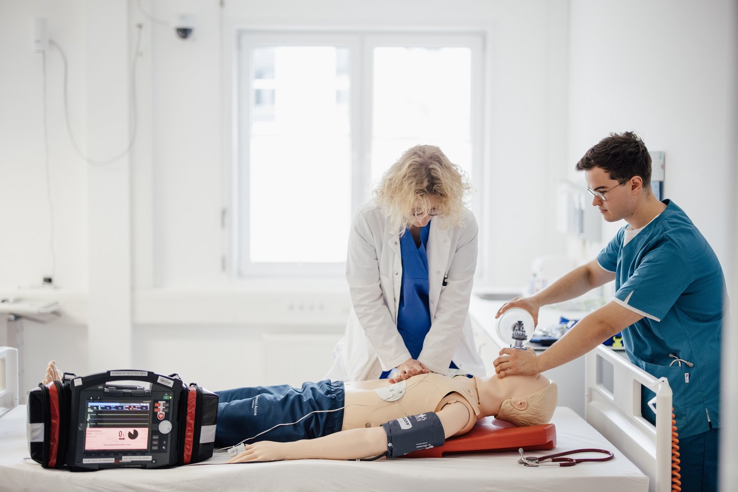 Eine medizinische Schulungssituation: Eine Person in einem weißen Kittel führt eine Herz-Lungen-Wiederbelebung an einer Übungspuppe durch, während eine weitere Person mit einem Defibrillator assistiert. Im Hintergrund ist ein heller Raum zu sehen.