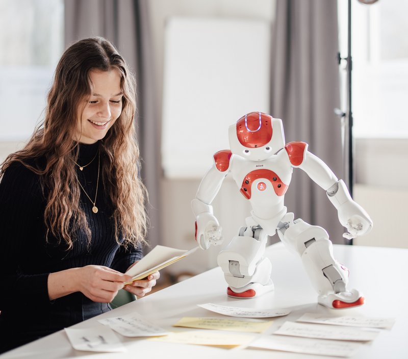 Eine Person sitzt an einem Tisch und betrachtet Karten, während ein humanoider Roboter daneben steht. Der Raum ist hell und modern, mit großen Fenstern und neutralen Farben.