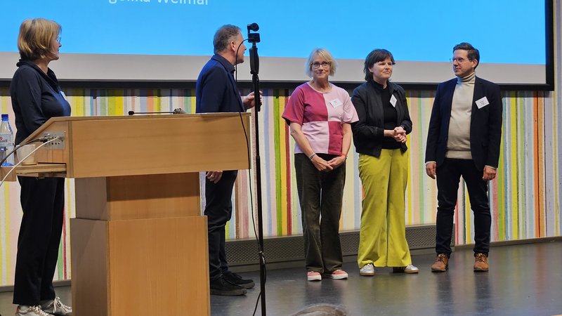 5 Personen stehen auf einer Bühne. Eine Person spricht.