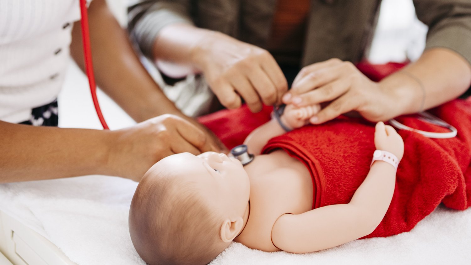 Eine Frau hört eine Babypuppe ab, welche in einer Neugeborenen-Reanimationseinheit liegt. Eine weitere Frau legt der Puppe einen Sensor an.