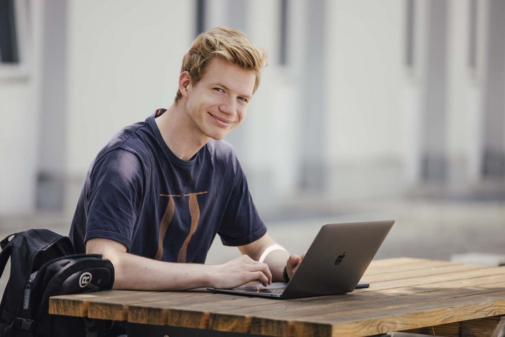 Eine Person sitzt draußen, lächelt und hat ihren Pc aufgeklappt.