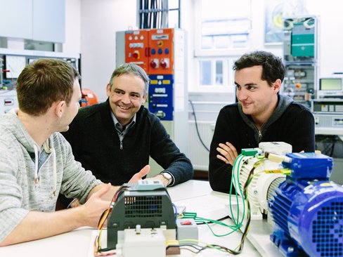 Ein Dozierender und zwei Studierende unterhalten sich. Vor ihnen steht ein Ausbau mit Motoren.