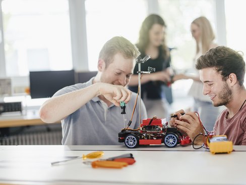 Zwei Personen schauen sich an und lächeln sie halten mit jeweils einer Hand einen Rover. Eine Person schraubt an dem Rover rum.