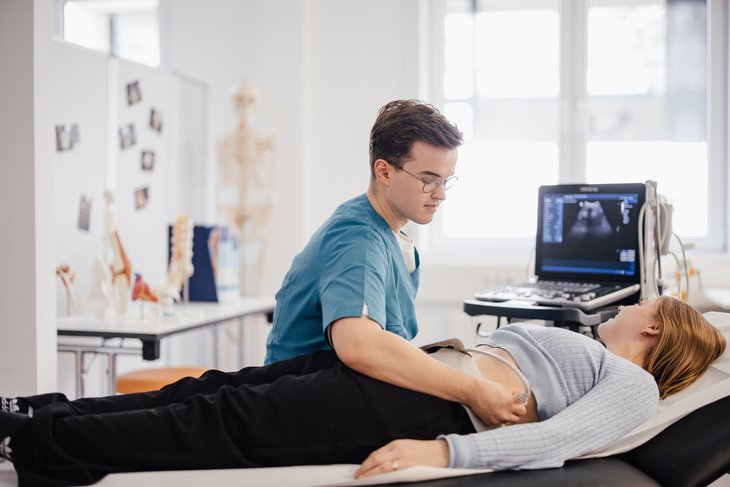 Eine Person in medizinischer Kleidung führt einen Ultraschalluntersuchung an einer liegenden Person durch. Im Hintergrund sind medizinische Modelle und ein Ultraschallgerät sichtbar.