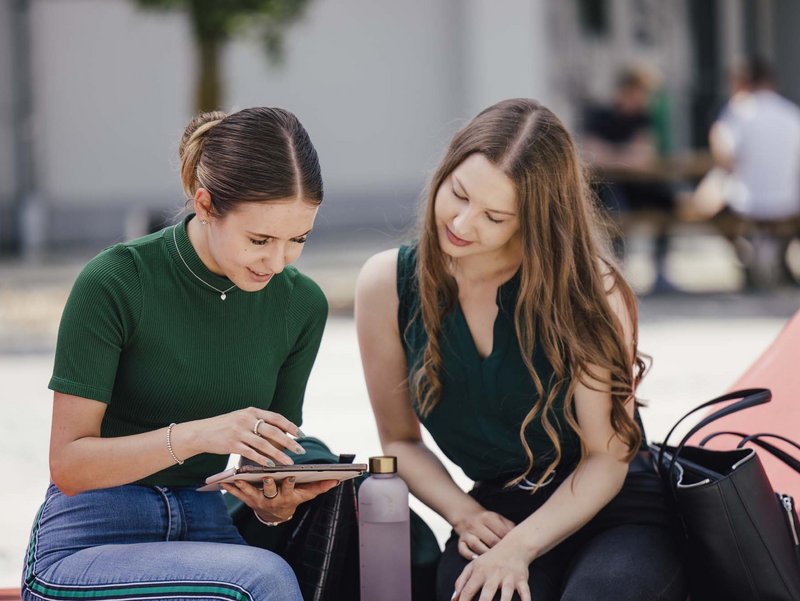 Zwei Personen sitzen draußen. Die eine hat ein ein Tablet in der Hand. Beide schauen ins Tablet.