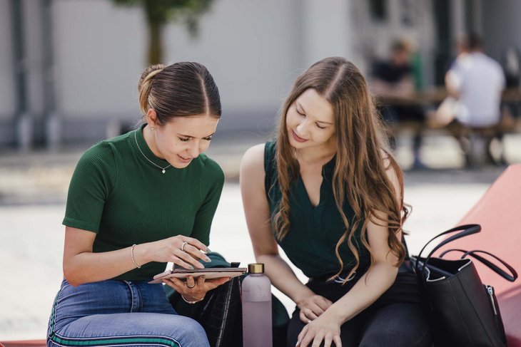 Zwei Personen sitzen draußen. Die eine hat ein ein Tablet in der Hand. Beide schauen ins Tablet.