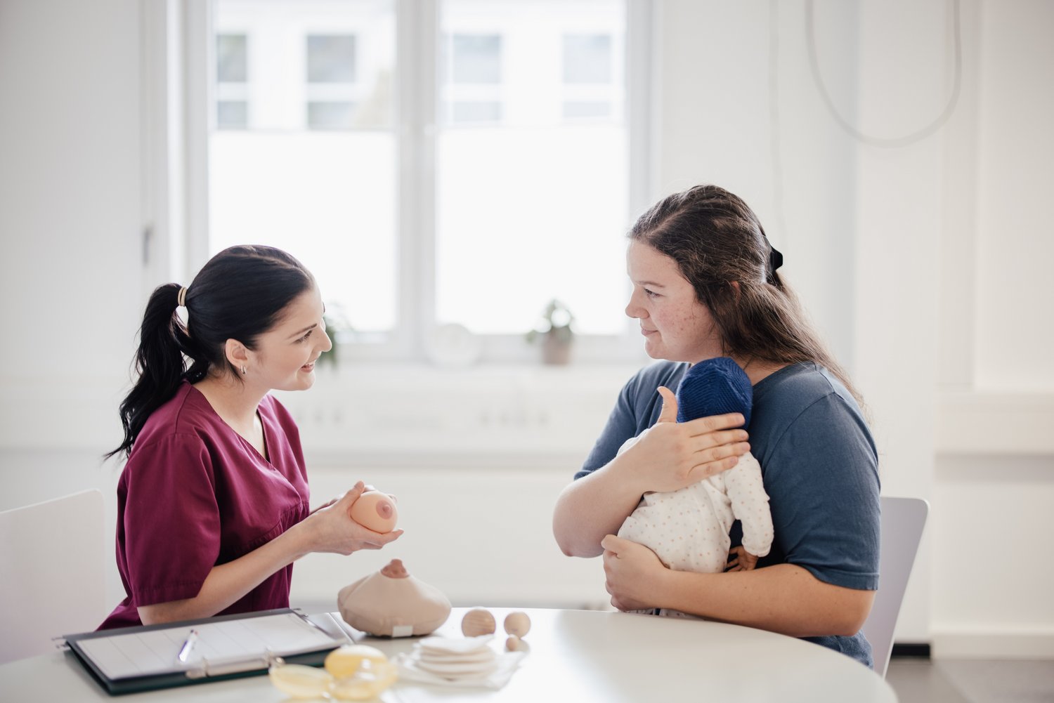 Eine Fachkraft erklärt einer Person mit einem Baby in den Armen etwas zu Stilltechniken. Auf dem Tisch sind verschiedene Hilfsmittel zur Stillberatung sichtbar. Der Raum ist hell und freundlich gestaltet.