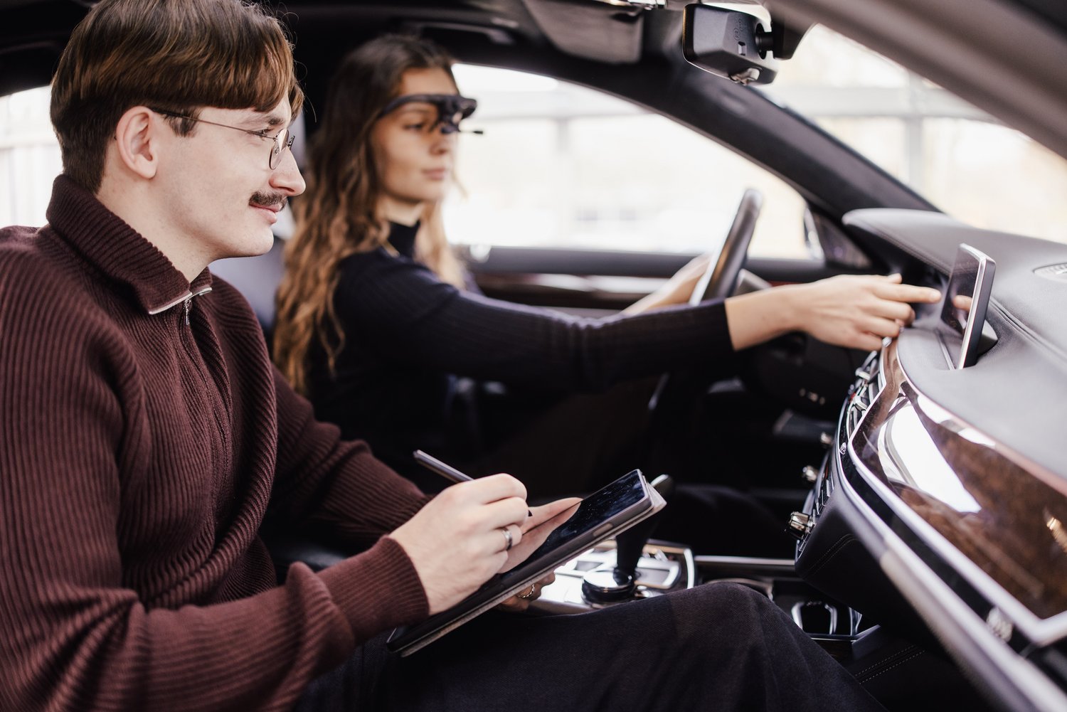 Eine Person sitzt auf dem Fahrersitz und bedient das Infotainmentsystem eines Autos, während eine andere Person auf dem Beifahrersitz Notizen auf einem Tablet macht. Beide sind in einem modernen Fahrzeug abgebildet.