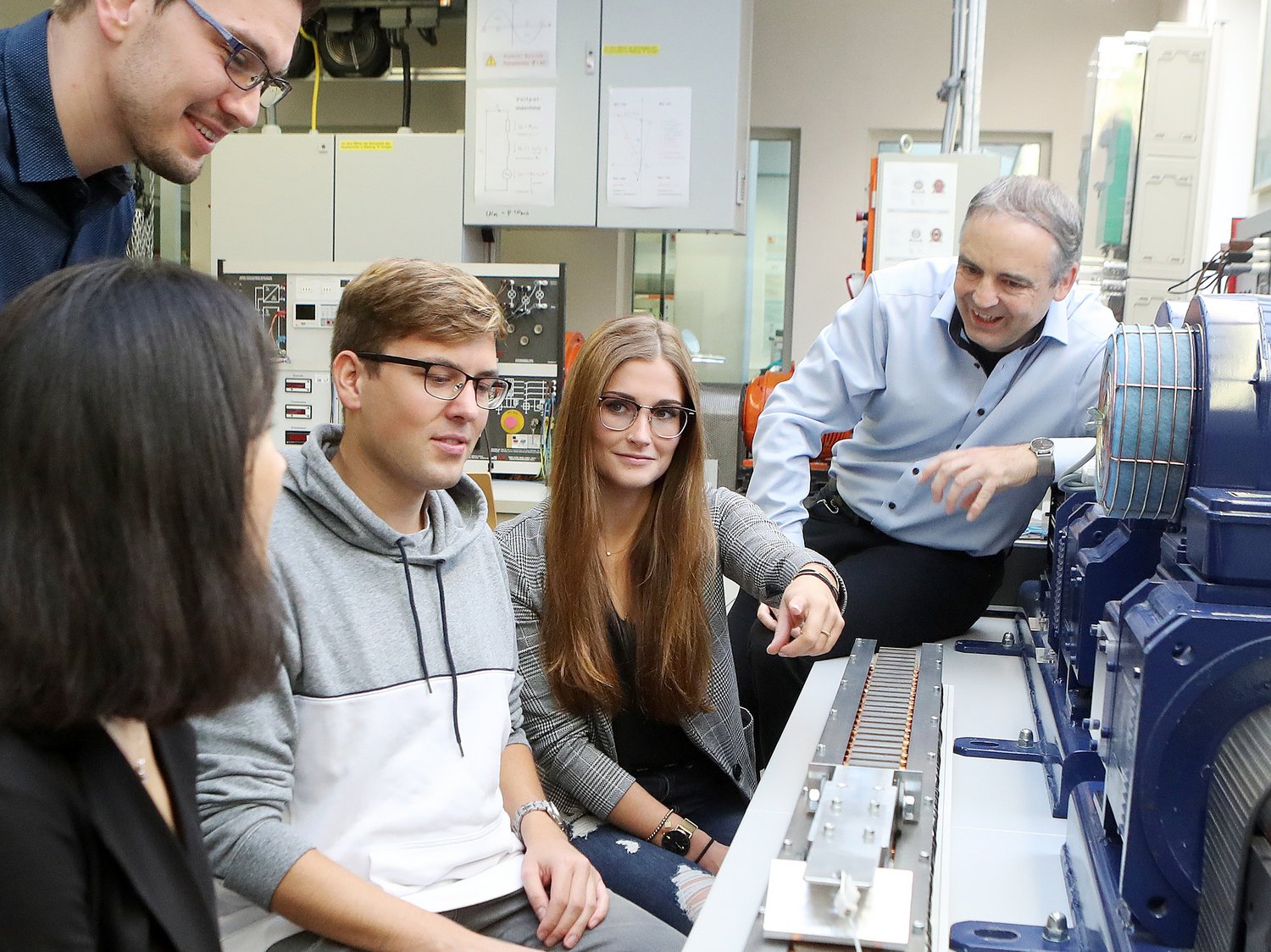 Mehrere Studierende und ein Professor sitzen vor dem Aufbau eines Linearmotors. Sie unterhalten sich.