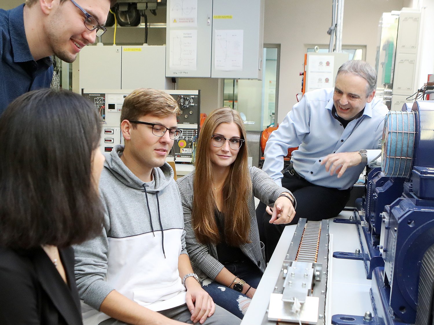 Mehrere Studierende und ein Professor sitzen vor dem Aufbau eines Linearmotors. Sie unterhalten sich.