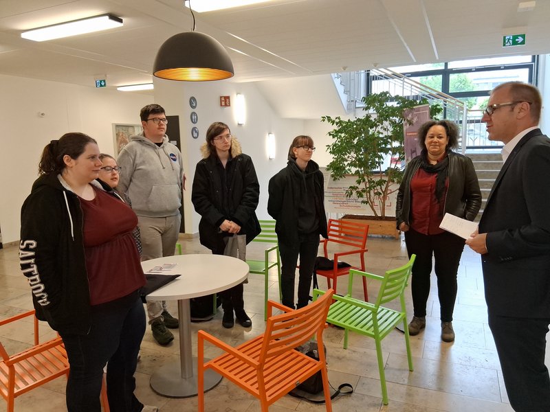 Gruppe Menschen steht vor einem Tisch und eine Person begrüßt sie.