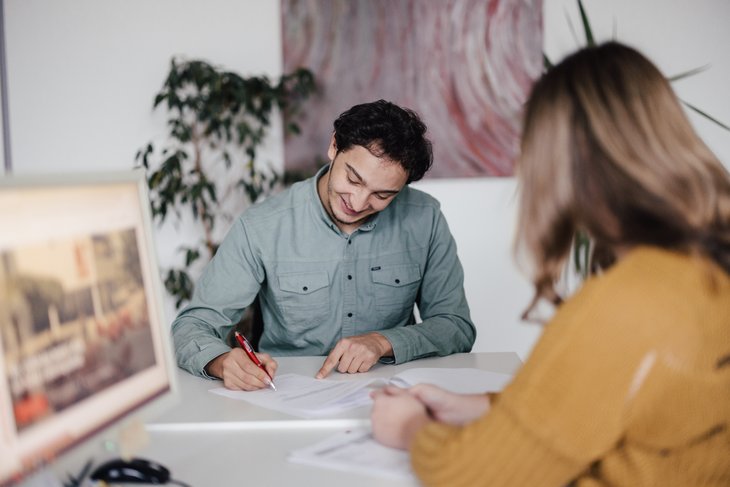 Zwei Personen sitzen an einem Tisch in einem modernen Büro. Eine Person schreibt Notizen, während die andere aufmerksam schaut. Im Hintergrund sind Pflanzen und ein Computerbildschirm sichtbar.