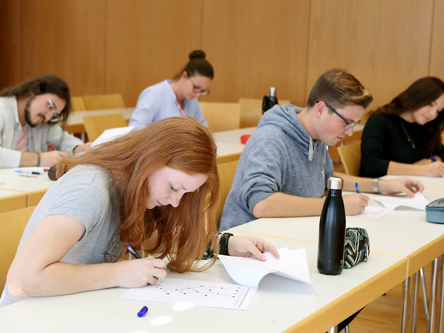 Fünf Personen sitzen in einem Vorlesungsraum und schreiben auf Blättern.