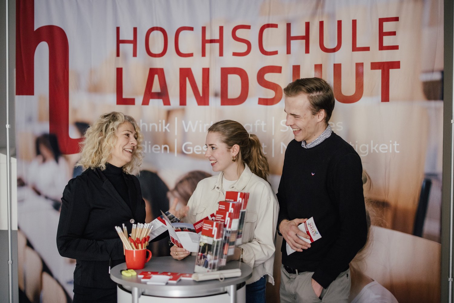 Drei Personen stehen an einem Informationsstand der Hochschule Landshut. Sie unterhalten sich und halten Broschüren in der Hand. Im Hintergrund ist ein großes Banner mit dem Schriftzug "Hochschule Landshut" zu sehen.