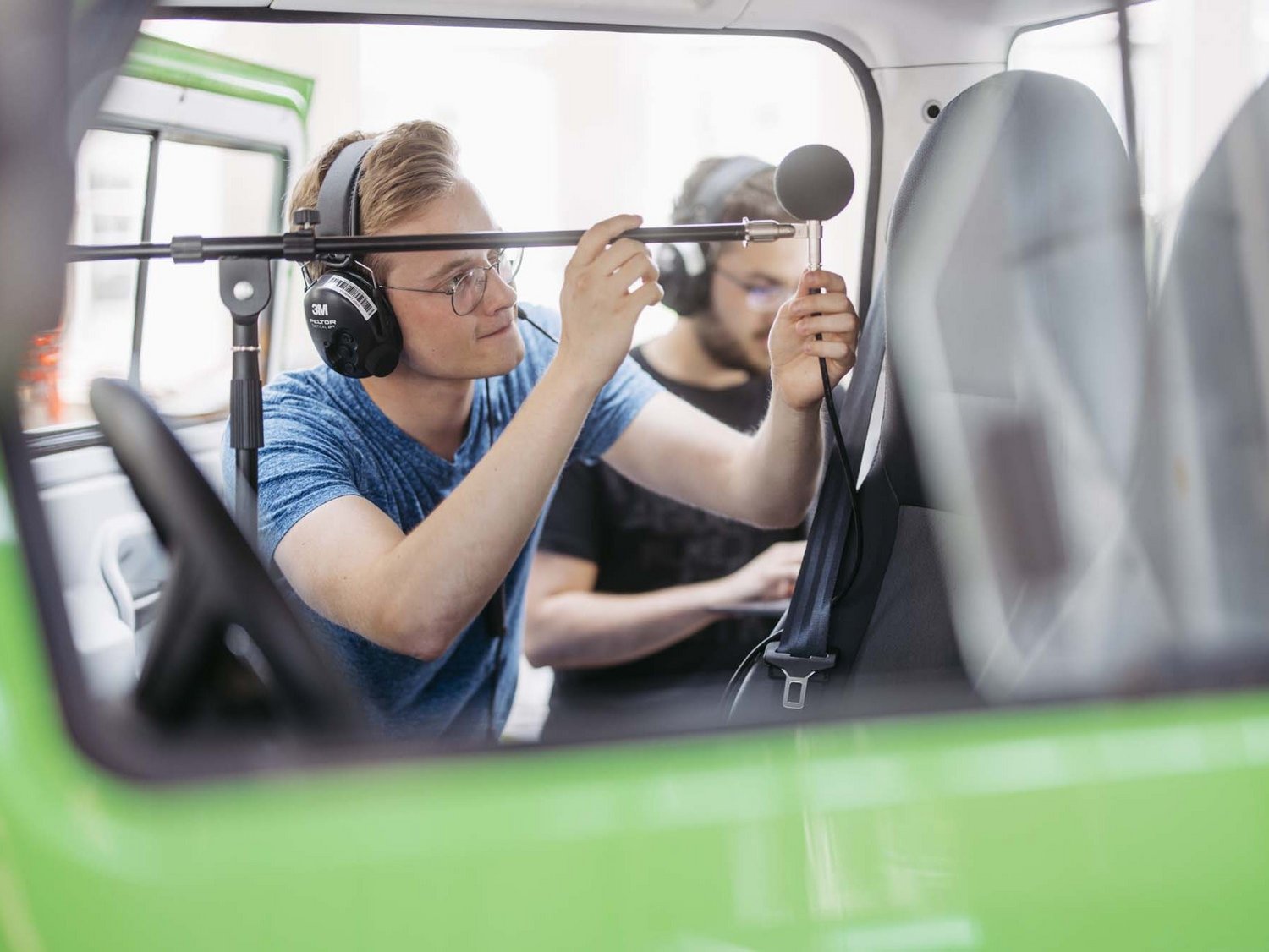 Zwei Studierende mit Köpfhörer bei der Schallpegelmessung mit einem Mikrophon auf einem Stativ in einer Fahrzeugkabine