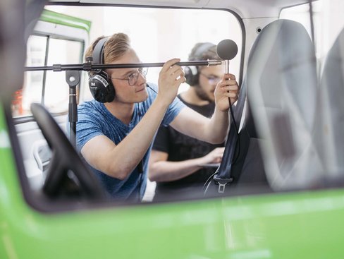 Zwei Studierende mit Köpfhörer bei der Schallpegelmessung mit einem Mikrophon auf einem Stativ in einer Fahrzeugkabine