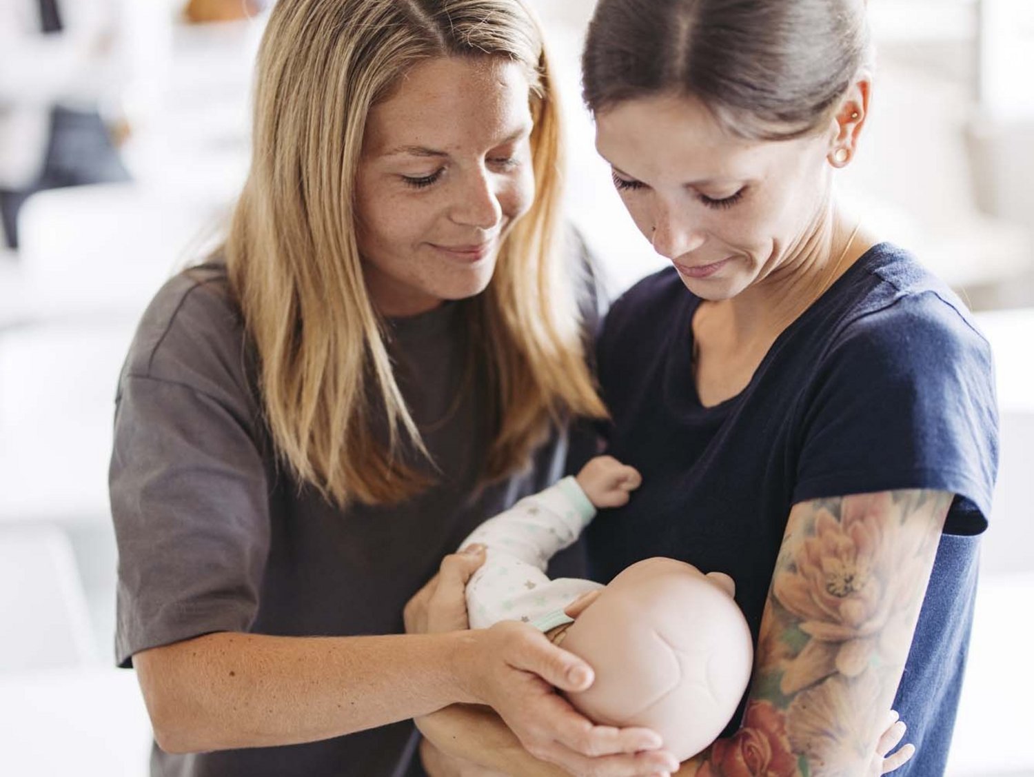 Zwei Personen sind zu sehen . Die eine hält eine Puppe die aussieht wie ein Baby in ihren Armen. Die andere Person schaut die Puppe an.