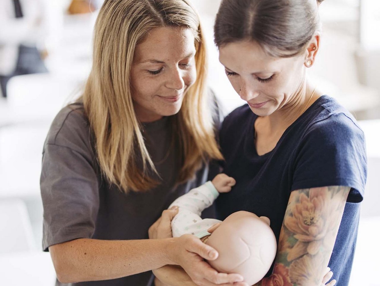 Zwei Personen sind zu sehen . Die eine hält eine Puppe die aussieht wie ein Baby in ihren Armen. Die andere Person schaut die Puppe an.