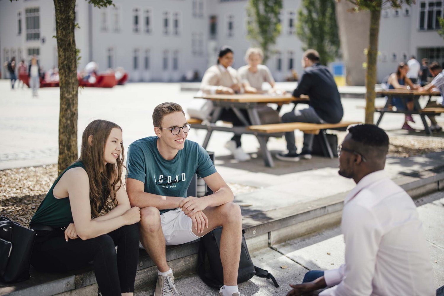 Drei Personen sitzen draußen. Die Sonne scheint. Sie unterhalten sich.