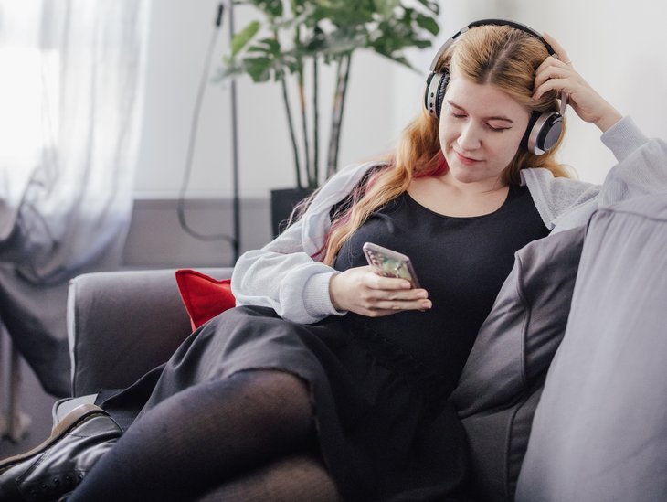 Eine Person sitzt entspannt auf einem grauen Sofa, trägt Kopfhörer und schaut auf ihr Smartphone. Im Hintergrund steht eine Pflanze und ein Stehlampe sorgt für sanftes Licht.
