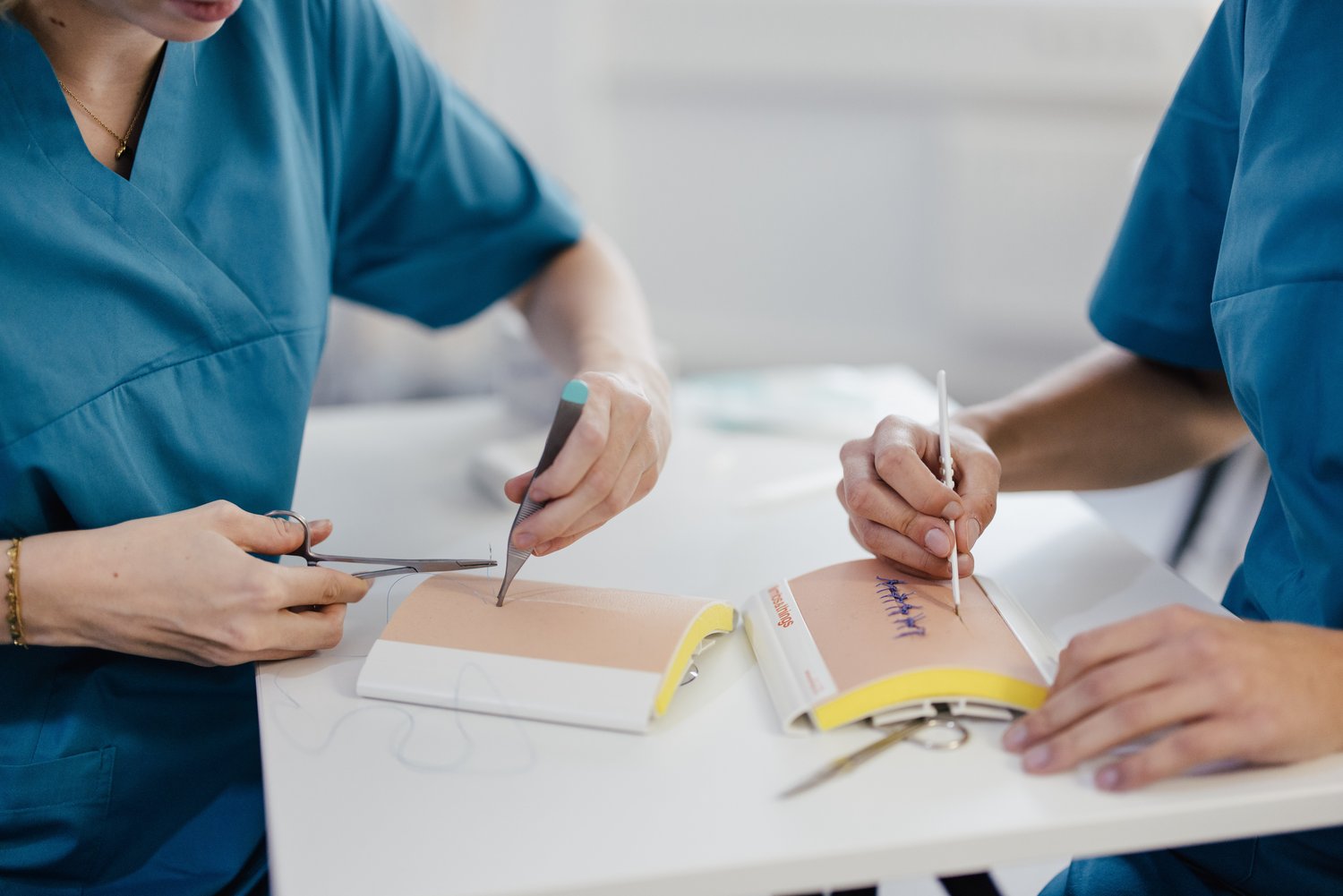 Zwei Personen in blauen medizinischen Kitteln arbeiten an einem Tisch. Eine Person schneidet mit einer Schere, während die andere mit einem Stift auf einem Modell schreibt. Im Hintergrund sind weitere medizinische Utensilien sichtbar.