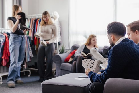 Eine Gruppe von fünf Personen in einem gemütlichen Raum. Zwei Personen stehen und halten Kleidung, während drei andere auf einem Sofa sitzen und Karten spielen. Im Hintergrund sind Kleiderständer und Fenster mit Tageslicht zu sehen.