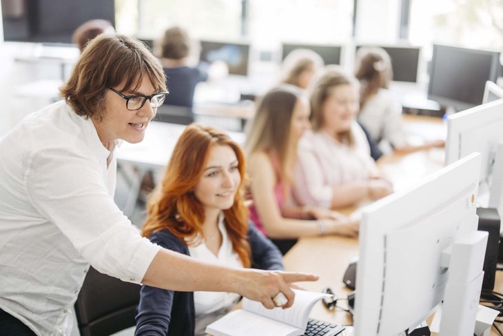 Zwei Personen sind in einem Raum. Die eine Person sitzt vor dem Computer, die andere Person steht dahinter und zeigt mit dem Finger auf dem Bildschirm..