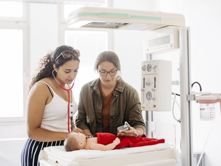 Zwei Frauen stehen an einer Neugeborenen-Reanimationseinheit in welchem eine Babypuppe liegt.