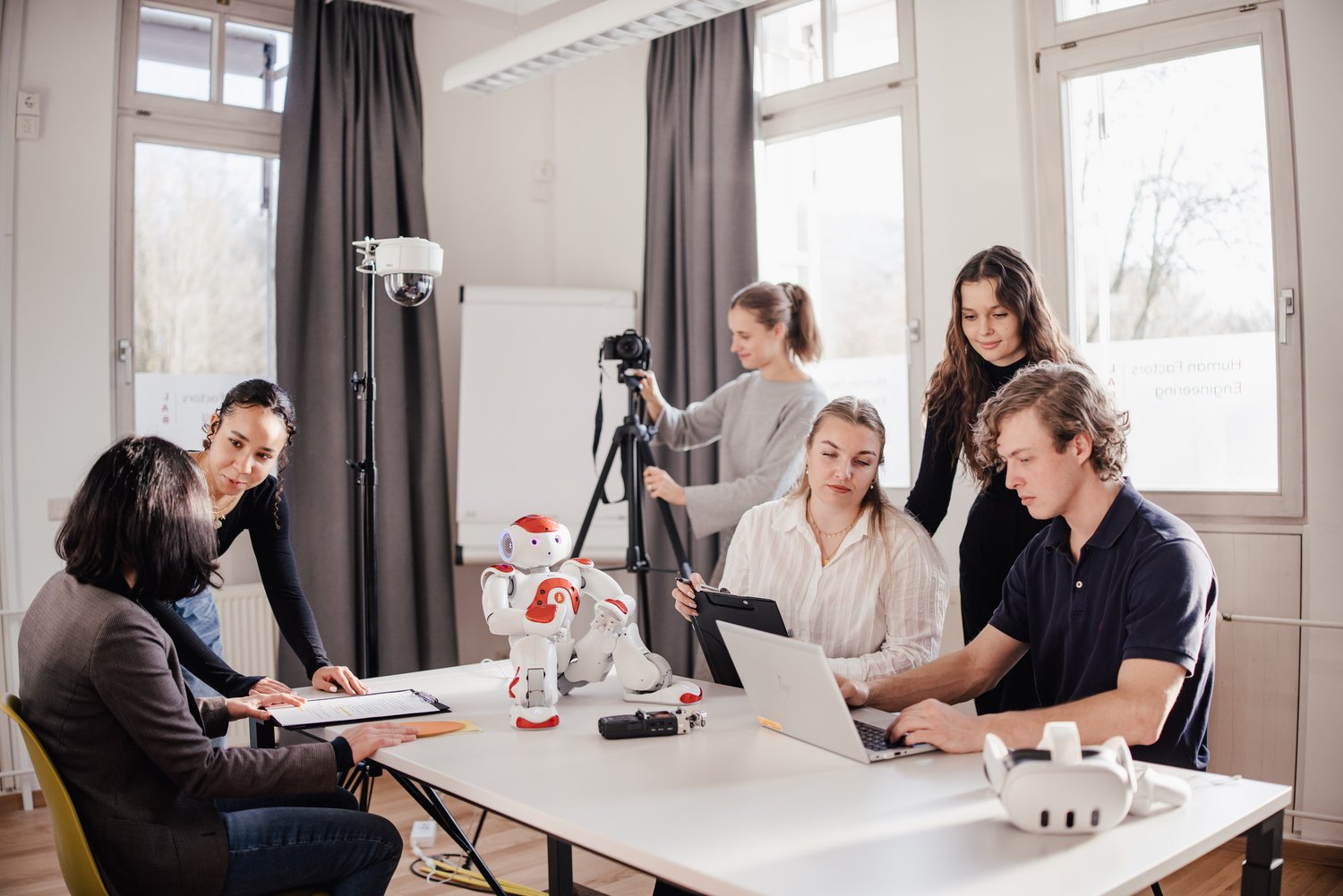 Eine Gruppe von Personen arbeitet in einem modernen Raum. Sie nutzen Laptops und Notizen, während ein humanoider Roboter und eine Kamera im Vordergrund stehen. Die Atmosphäre ist kreativ und kollaborativ.