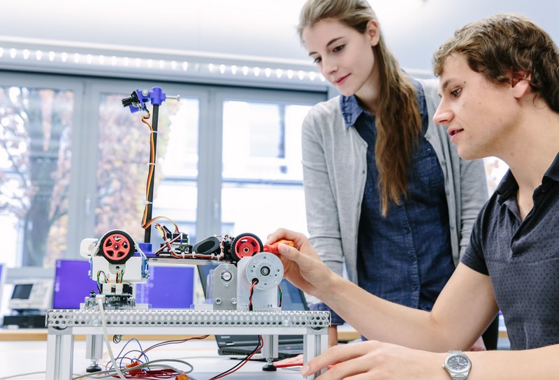 Zwei Studierende nehmen ein kleines autonomes Fahrzeug auf einem Teststand in Betrieb.