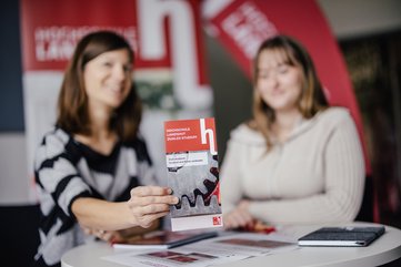 Zwei Personen sitzen an einem Tisch und halten ein Informationsblatt über ein Hochschulangebot. Im Hintergrund sind rote Banner sichtbar. Die Atmosphäre wirkt freundlich und einladend.